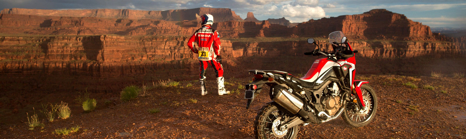 A 2020 red Honda® CRF250L parked next to the canyon's edge with the rider gazing off into the distan…